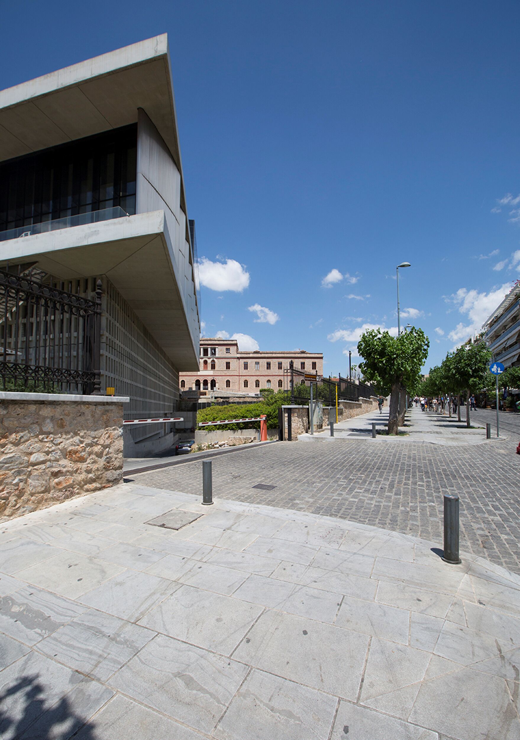 A paved pedestrian area with a modern building, stone wall, and a tree.