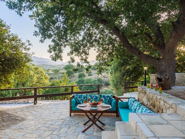 A stone patio with a wooden table and blue sofa, surrounded by trees and a stone wall.