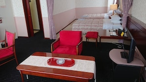 A hotel room with a bed, a desk with a tray, two red chairs, a television, and a window with curtains. - Mihara Kokusai Hotel (Mihara)