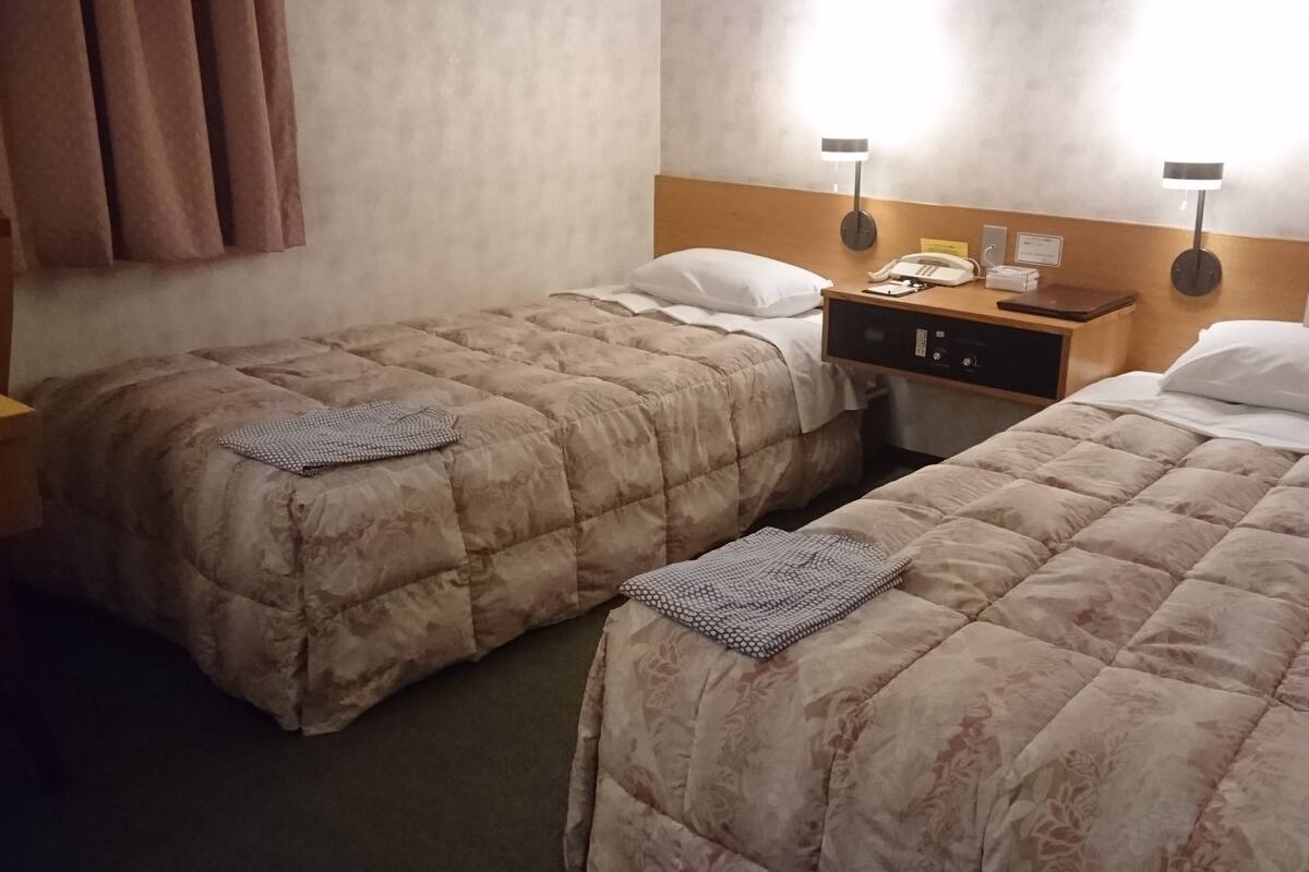A hotel room with two beds, each with a quilted duvet and pillows, a bedside table with a lamp, a phone, and a small card holder.