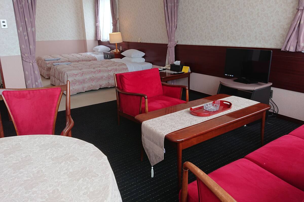 A hotel room with a bed, two chairs, a table with a tray, a TV, and a desk with a lamp.