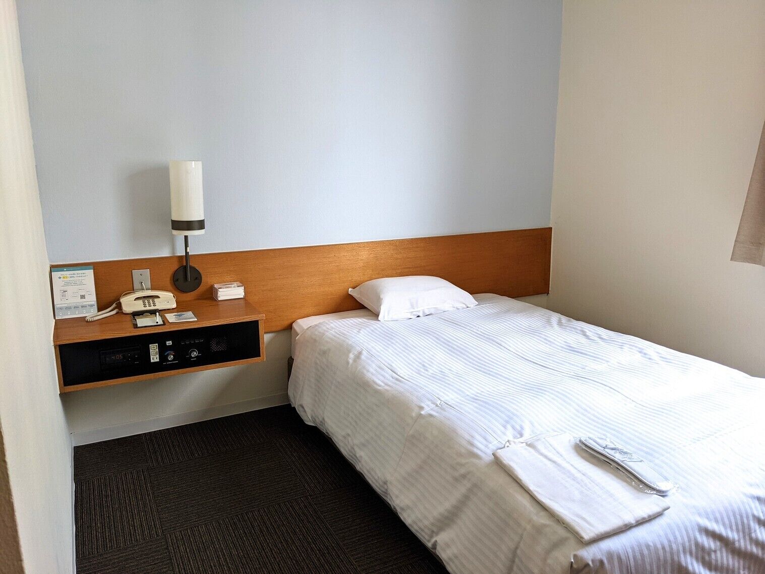 A hotel room with a bed, a bedside table with a lamp, a phone, and a control panel.