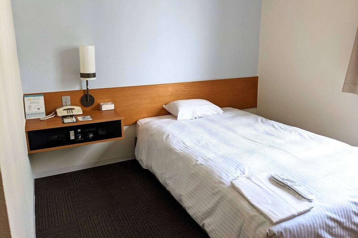 A hotel room with a bed, a bedside table with a lamp, a phone, and a control panel.