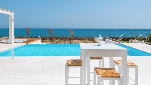 Un área junto a la piscina con una mesa y sillas blancas, vista despejada al mar y una pared de piedra.