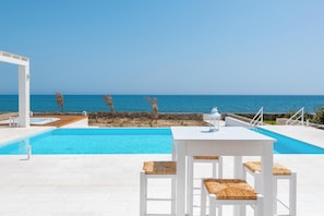 A poolside area with a white table and chairs, a clear view of the sea, and a stone wall.