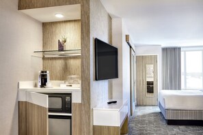 Quarto de hotel moderno com cozinha compacta, televisão de tela plana, cama e uma grande janela.