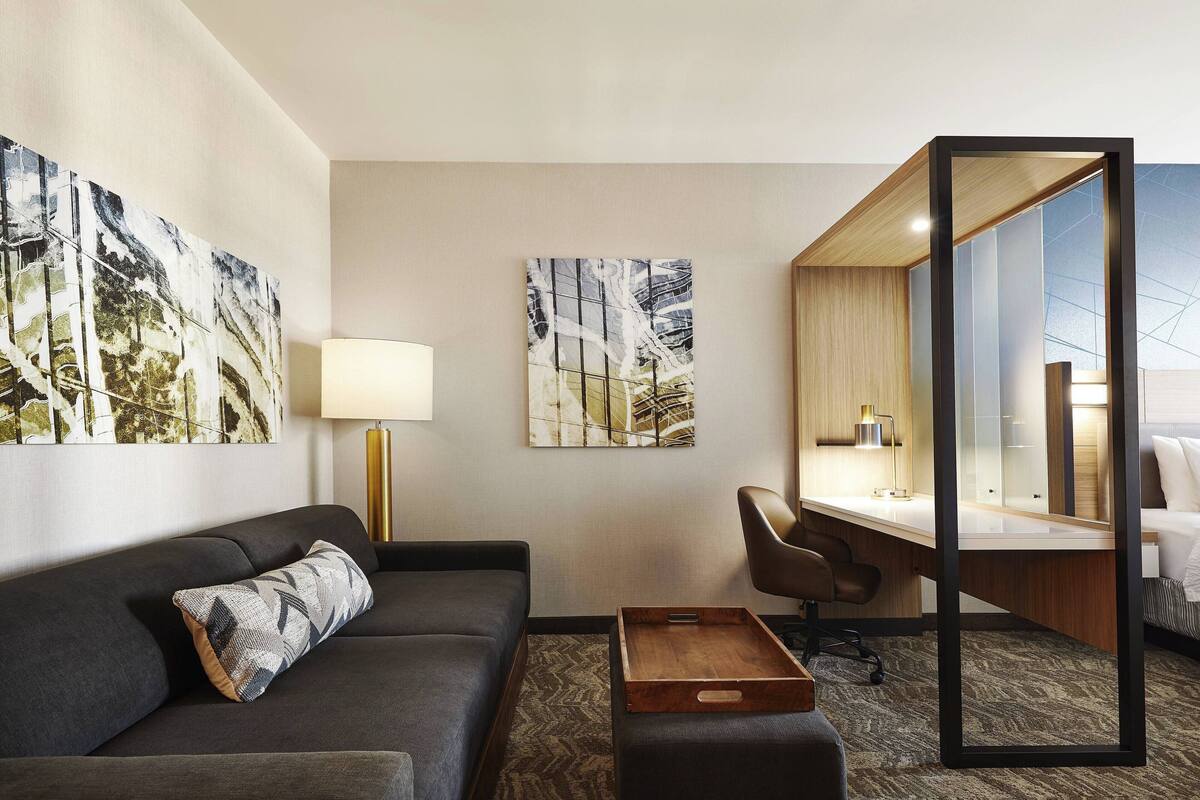 A modern hotel room with a dark grey sofa, a wooden side table, a desk with a chair, and two abstract paintings on the walls.
