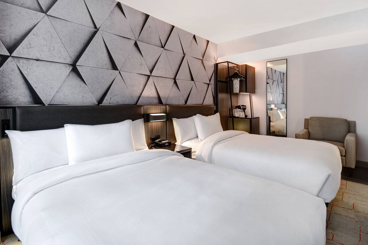 A hotel room with two beds, a modern geometric headboard, a bedside table with a lamp, a chair, and a large mirror.