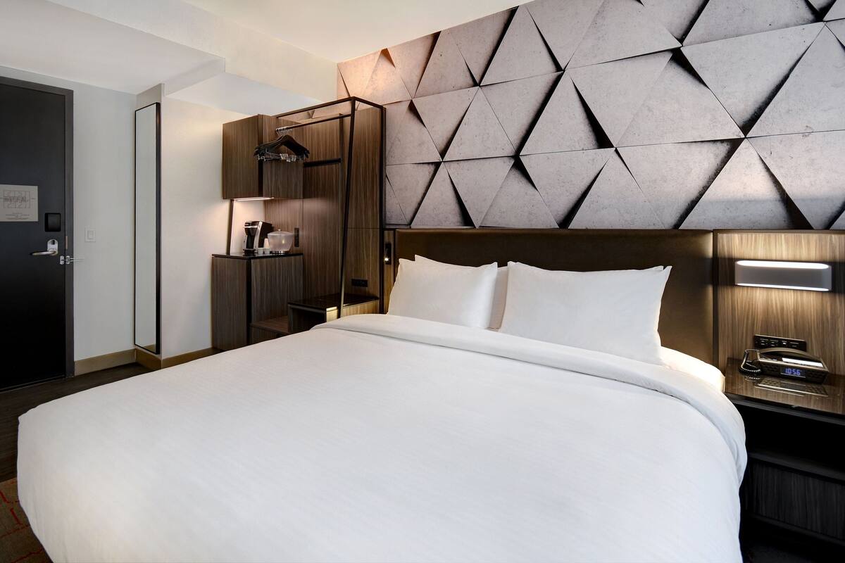 A modern hotel room with a large bed, a wooden headboard, a built-in wardrobe, and a bedside table with a lamp and alarm clock.