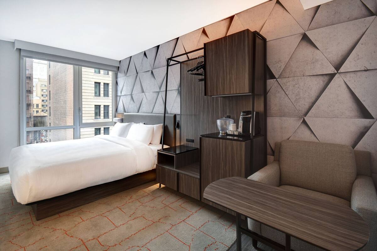 A modern hotel room with a large bed, a sofa, a desk, and a wardrobe. There is a window providing a view of the city.