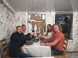 En gruppe mennesker sidder omkring et bord med hvid dug, med vinglas i hånden, i et rum med tapet og en trædør.