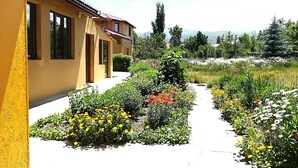 Un bâtiment jaune avec un toit en tuiles, un jardin où poussent différentes sortes de fleurs et un ciel dégagé.
