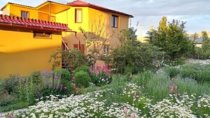 Une maison jaune avec un toit rouge, entourée d’un jardin où poussent différentes sortes de fleurs et de plantes.