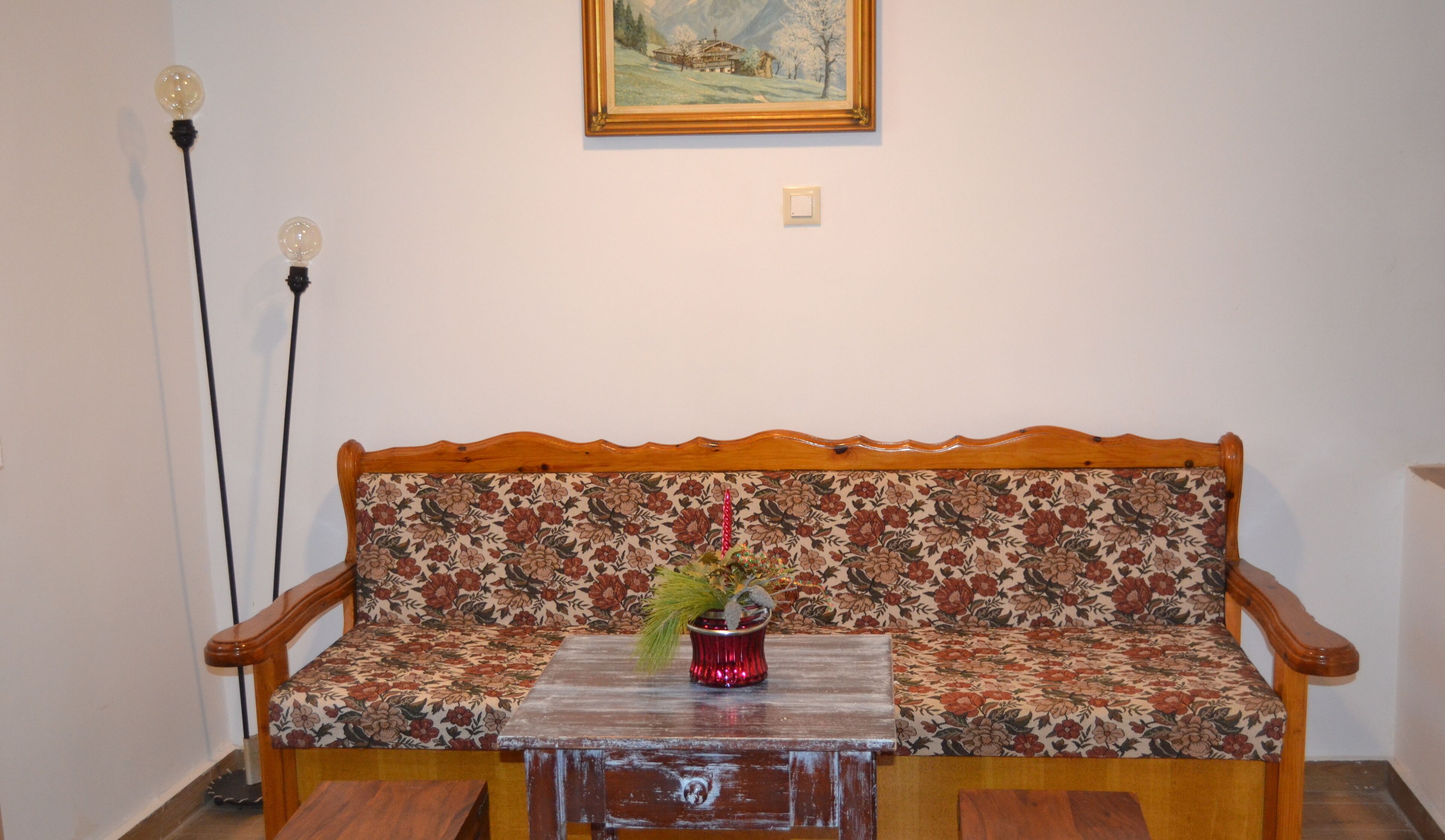 A wooden sofa with floral upholstery, a small table with a red vase, and a painting on the wall.