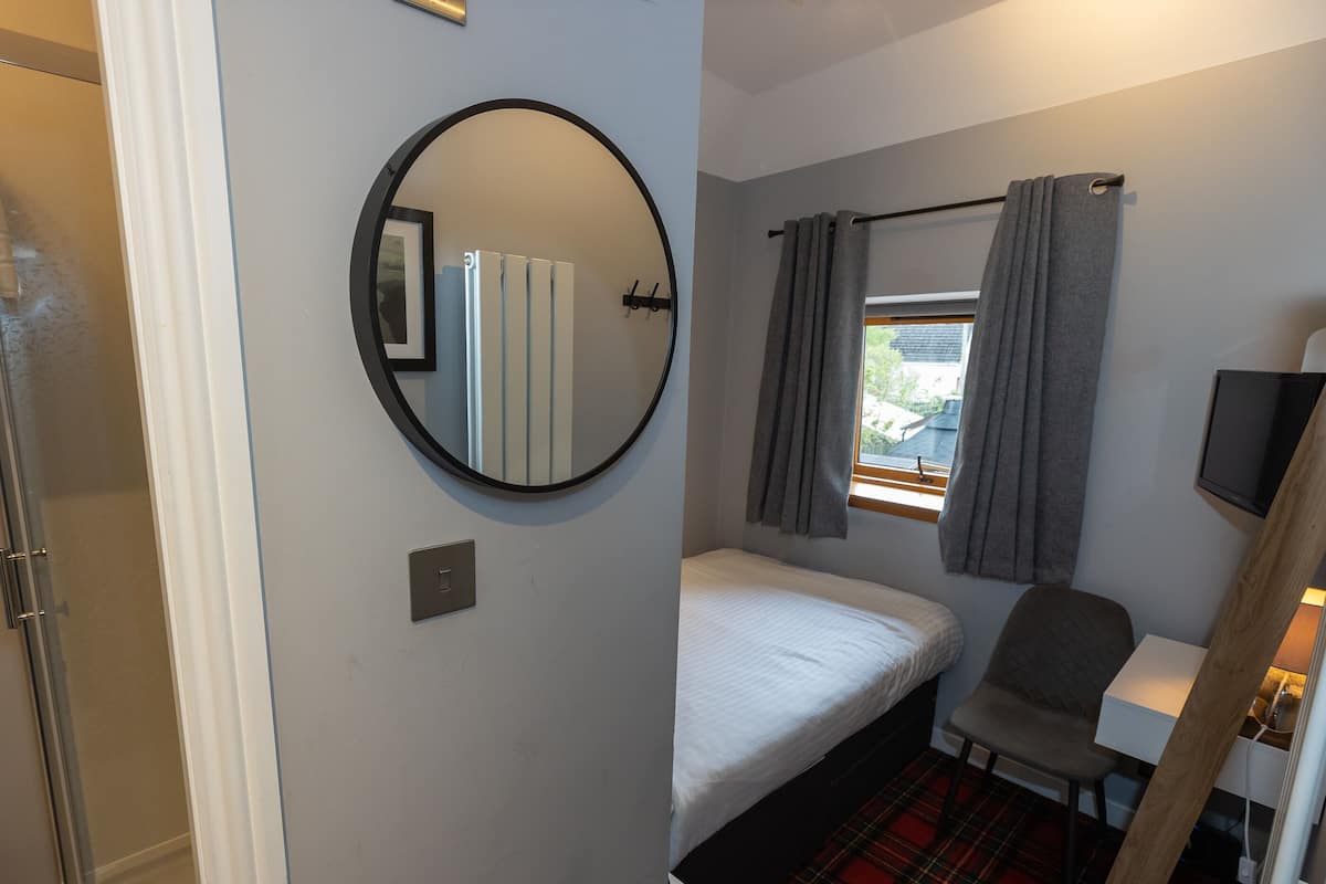 Habitación de hotel compacta con una cama individual, un espejo redondo, una mesita pequeña, una silla, un televisor y una ventana con cortinas.