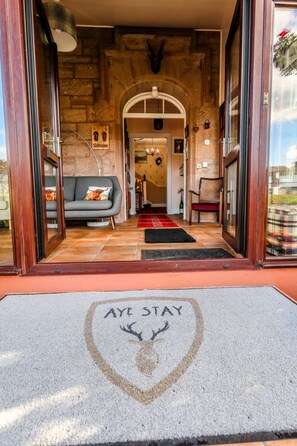 Entrada de hotel con fachada de piedra, un sofá con cojines, una alfombra roja y negra, y un felpudo con la inscripción “AYE STAY” y la silueta de un ciervo.
