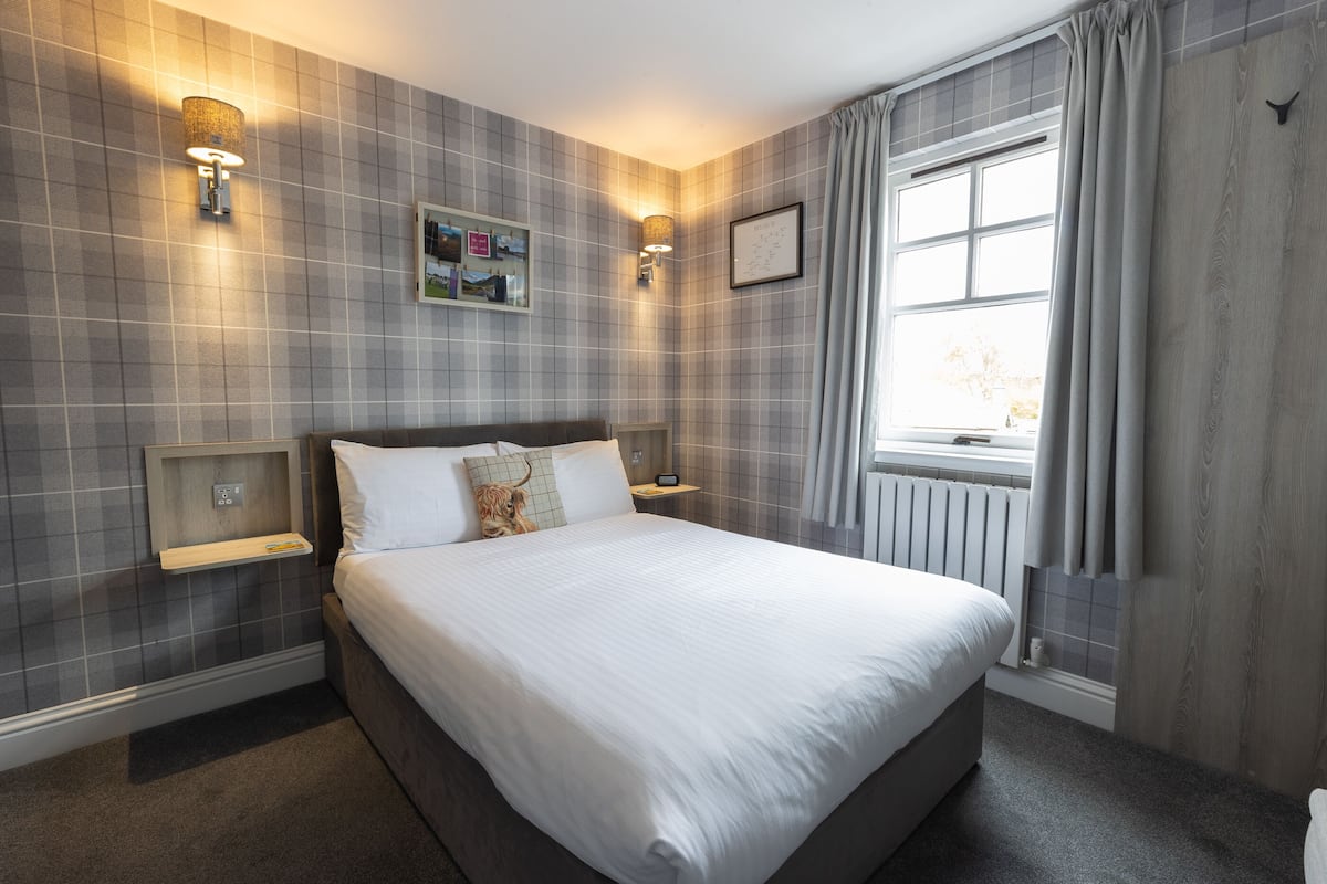 Una cama bien hecha en una habitación de hotel con un patrón de pared a cuadros, dos mesitas de noche con lámparas, una ventana con cortinas y un radiador.
