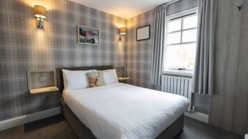 A neatly made bed in a hotel room with a checkered wall pattern, two bedside tables with lamps, a window with curtains, and a radiator.