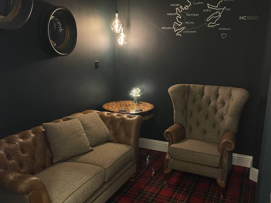 Un salón con decoración oscura, que incluye un sofá de cuero, un sillón capitonado, una alfombra a cuadros y un mapa mural.