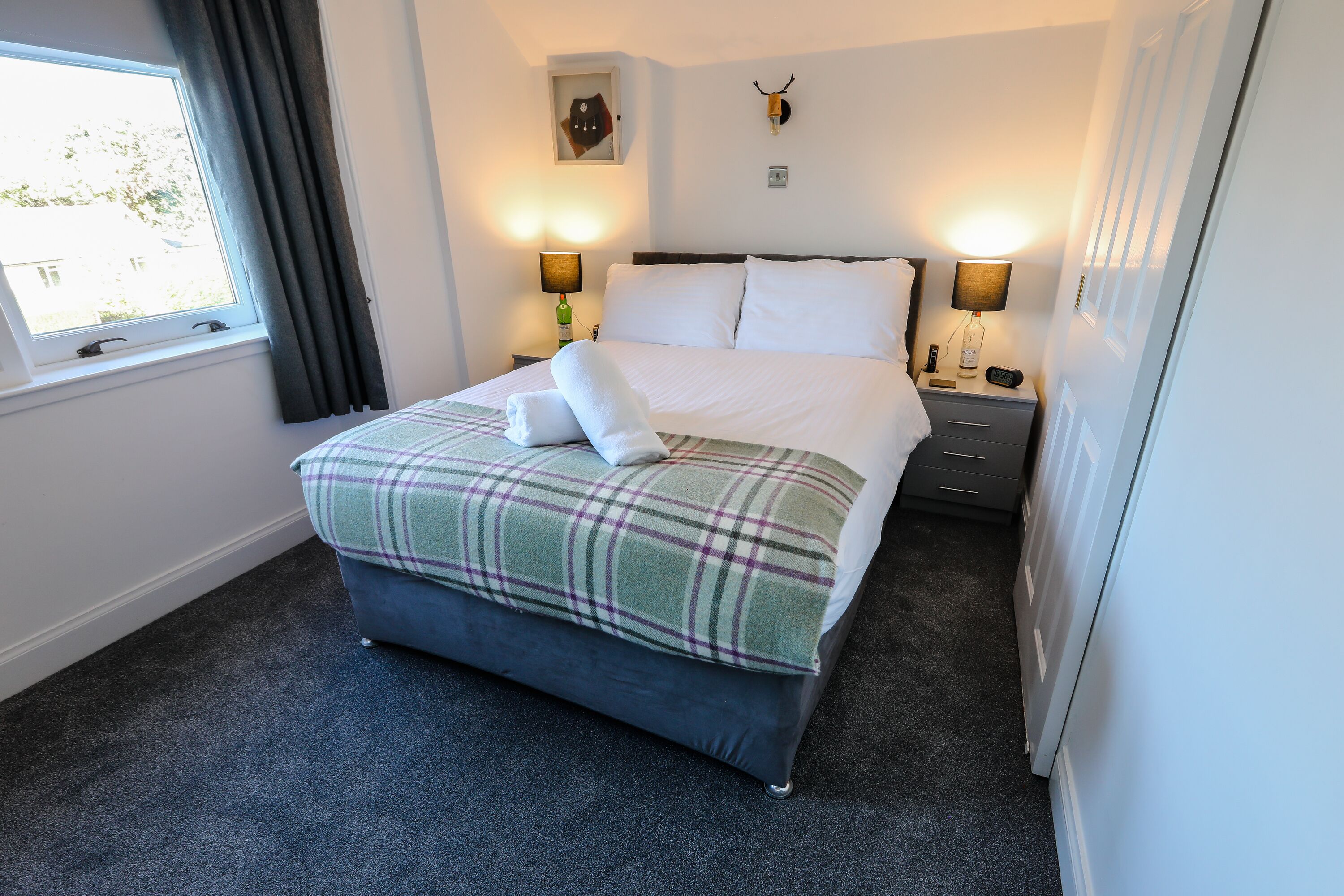 A neatly made bed with a plaid blanket and a folded towel, two bedside lamps, a small table with a lamp, and a wall-mounted deer head decoration.