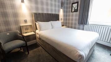 A hotel room with a large bed, a nightstand, a chair, a wall-mounted light, and a window with curtains.