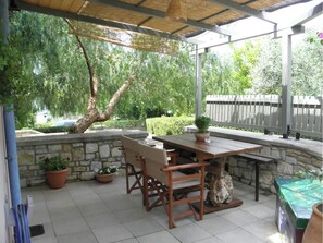 En udendørs patio med stenvæg, træbord og stole, omgivet af grønne planter og et træhegn.