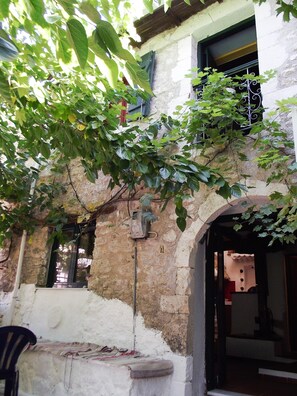Un edificio de piedra rústico con un pequeño balcón, una ventana con reja de metal y un árbol de hojas verdes que parcialmente bloquea la vista.