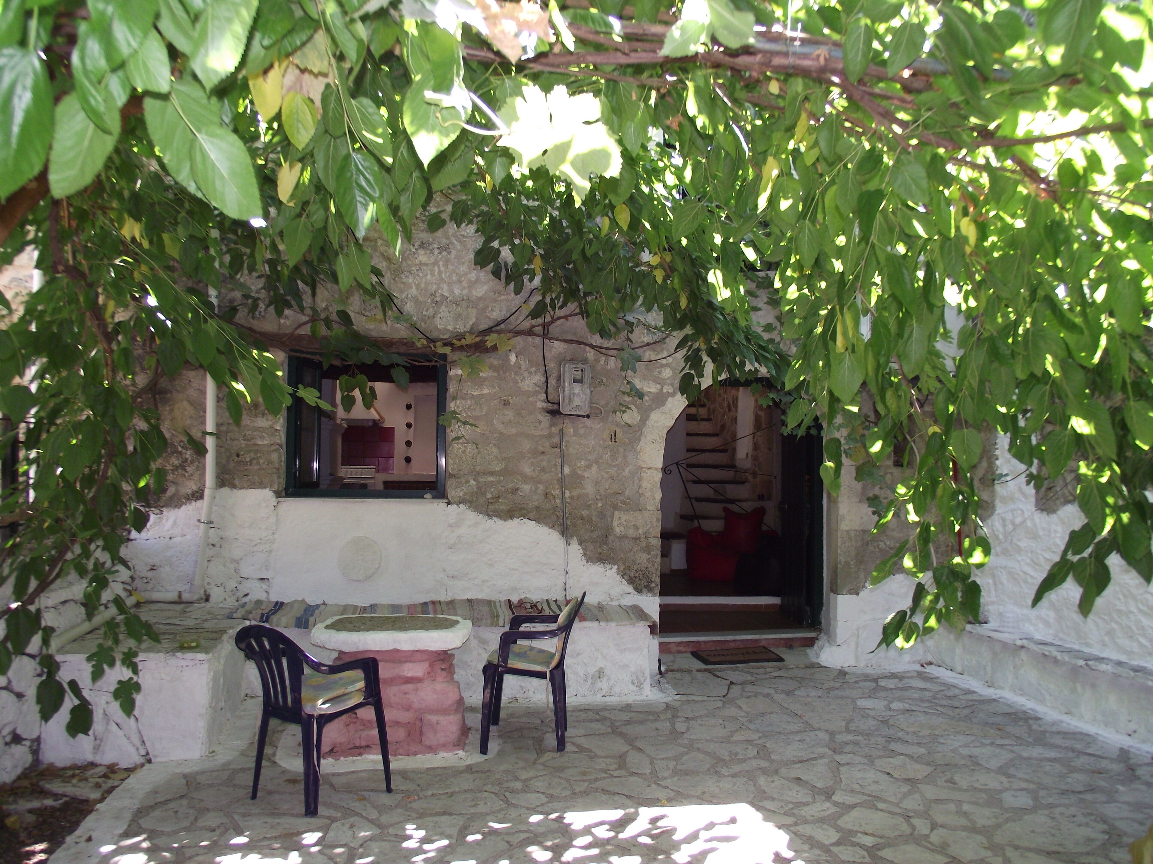 A stone courtyard with a rustic stone bench and two chairs, a small table, and a doorway leading to an interior space.