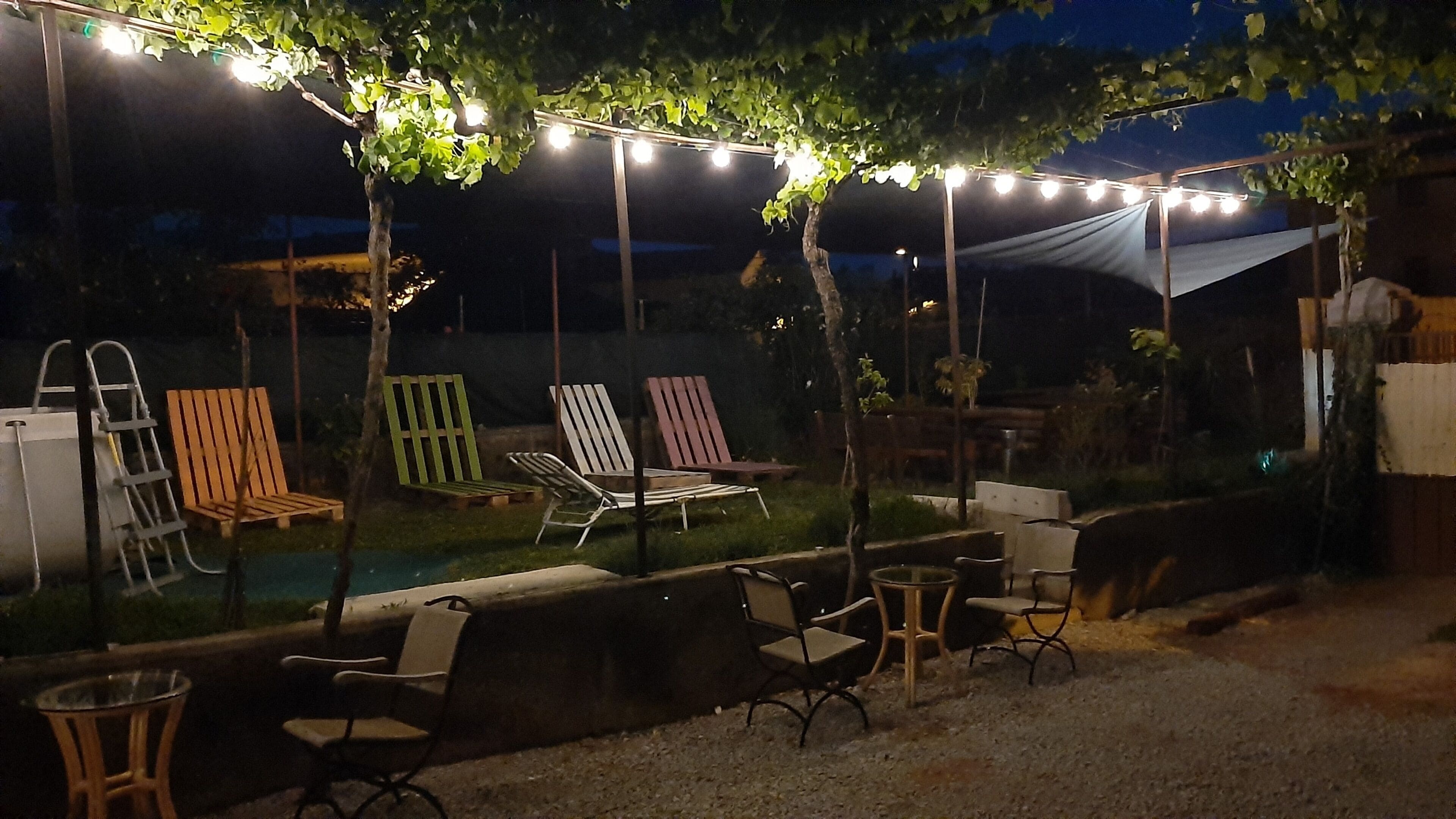 An outdoor patio area with string lights, wooden chairs, a pool, and a covered seating area.