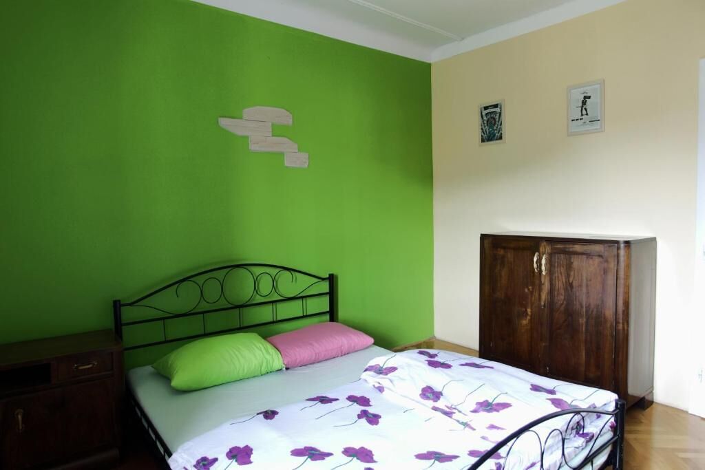 A bedroom with a green wall, a bed with a floral bedspread, a wooden wardrobe, and a small nightstand.