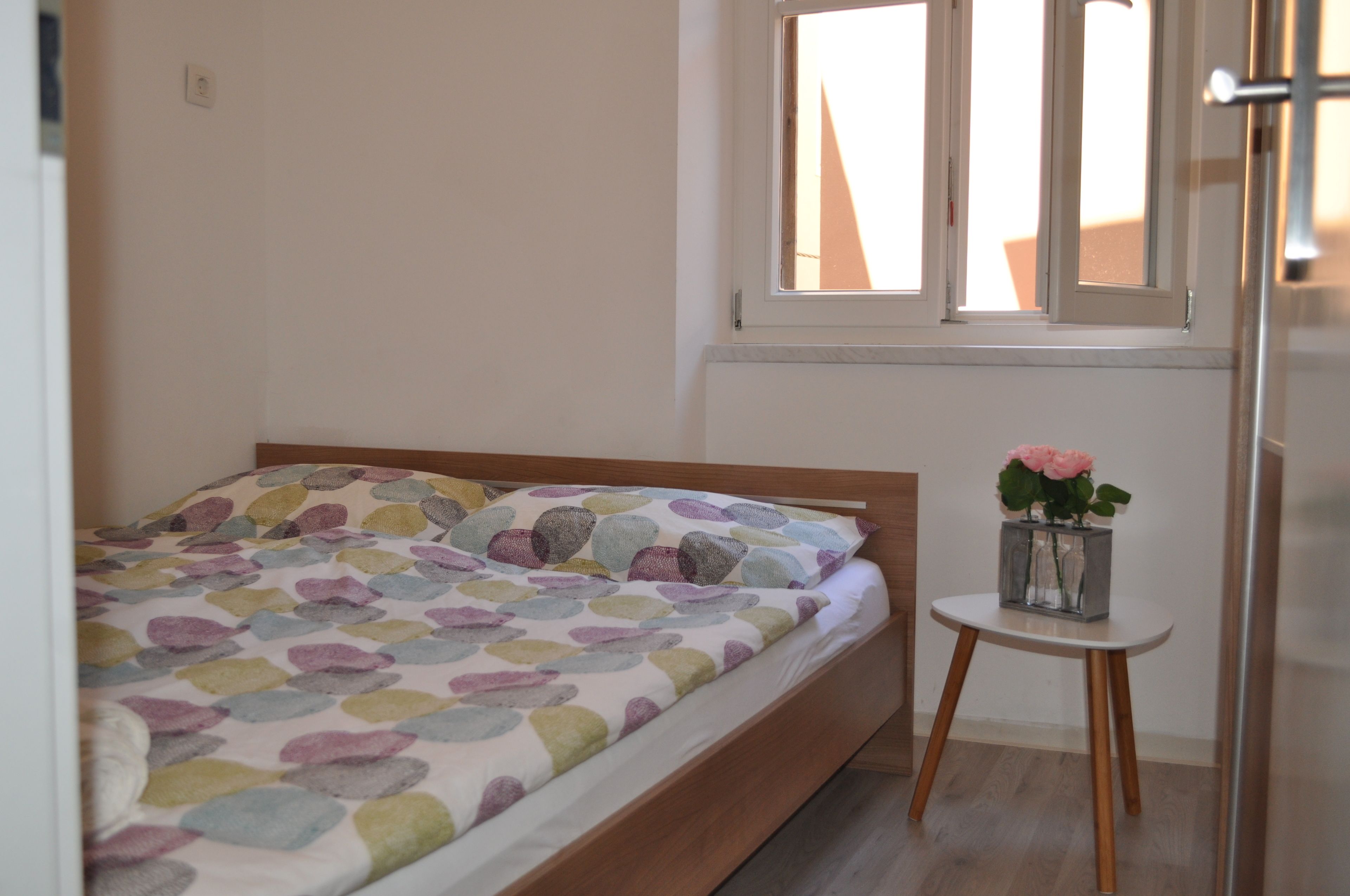 Un dormitorio con una cama, una mesita con un jarrón de flores y una ventana con vistas al exterior.