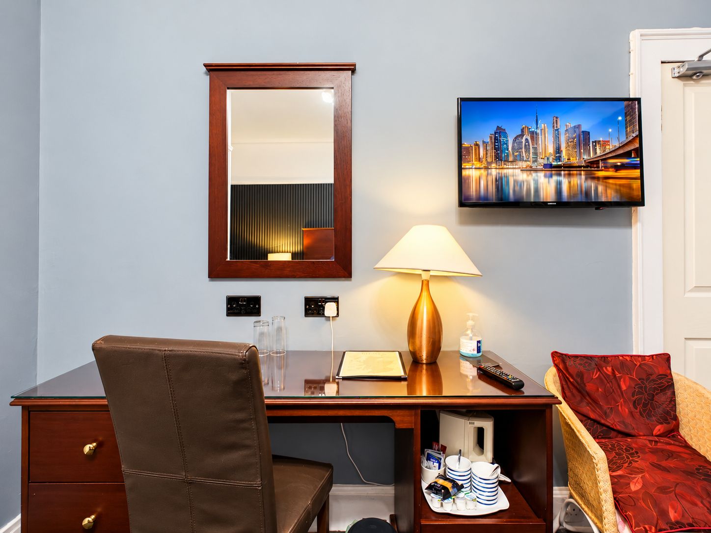 Een hotelkamer met een bureau, stoel, nachtkastje, lamp, afstandsbediening en een televisie waarop een stadsgezicht te zien is.