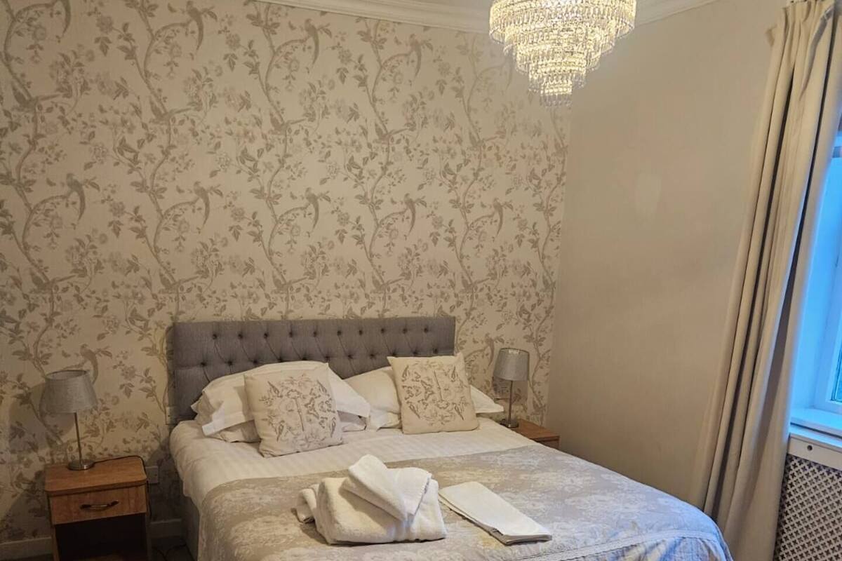 A neatly made bed with a grey headboard, two bedside tables with lamps, floral wallpaper, and a large window with curtains.
