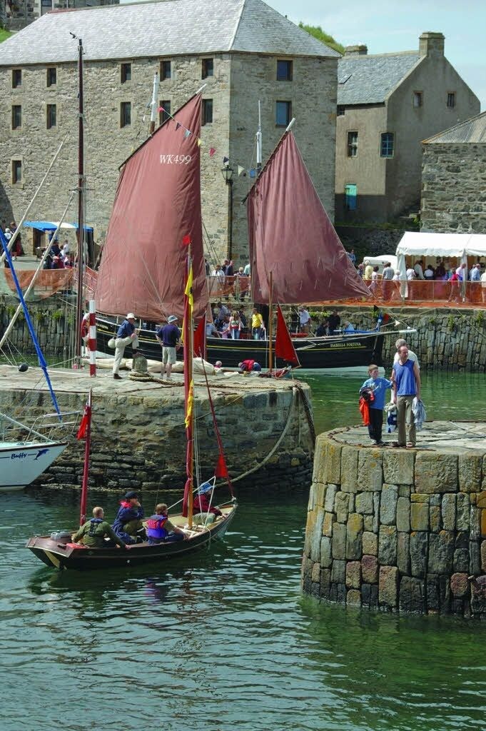 A stone pier with a sailboat docked, a building in the background, and a person standing on the pier.