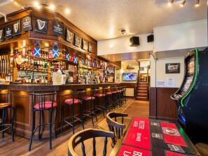 Zona de bar con taburetes, una barra con varias botellas, banderas y una pantalla de televisión.