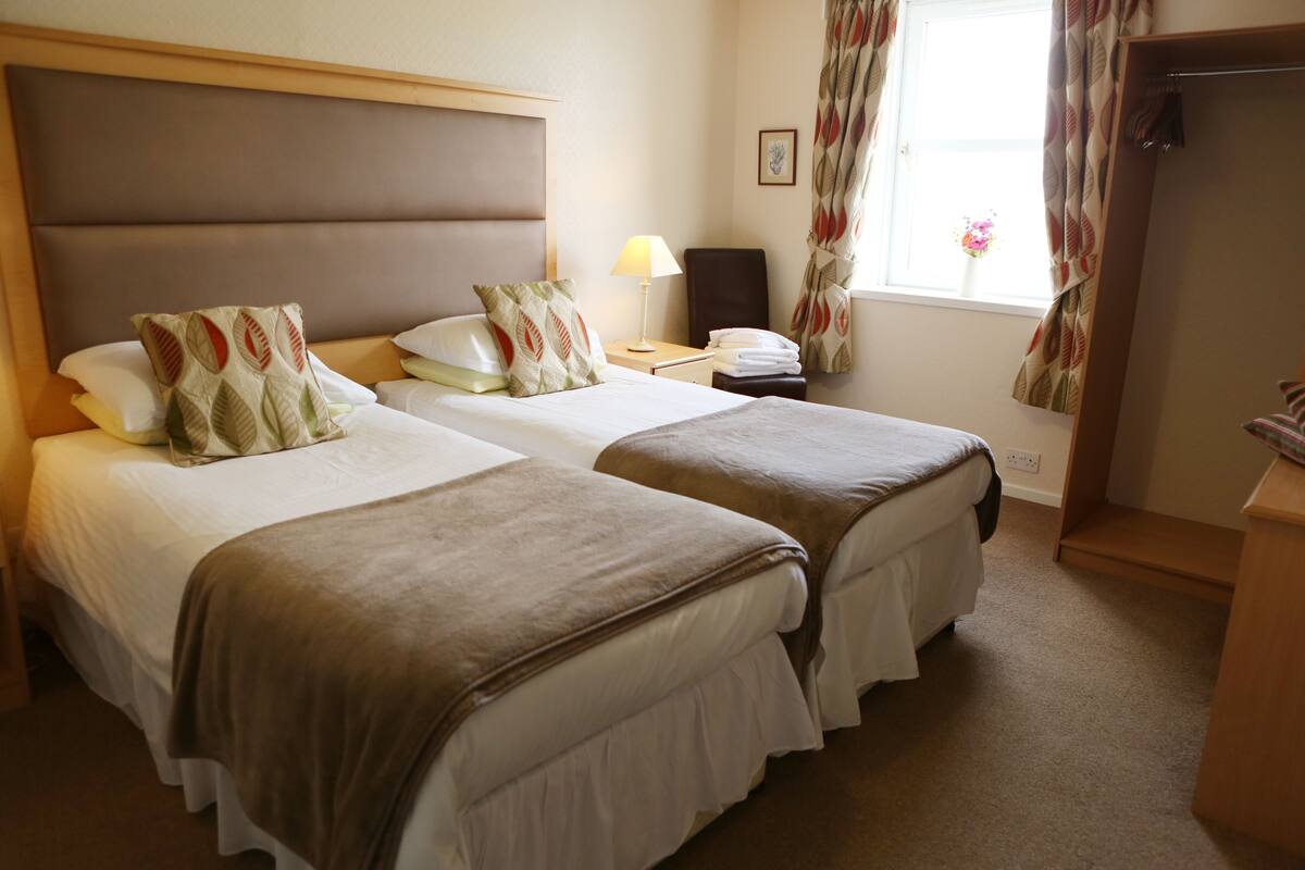 A hotel room with two beds, a desk, a chair, a lamp, and a window with curtains.
