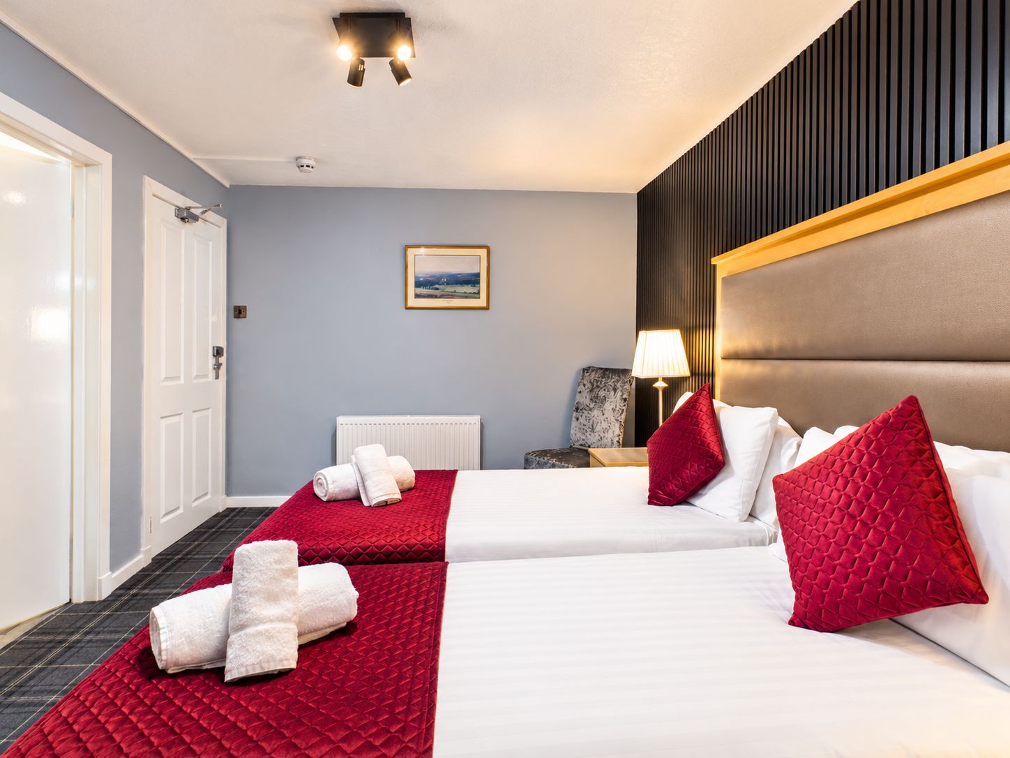 Une chambre d’hôtel avec deux lits, une couvre-lit rouge matelassée, du linge de lit blanc, un oreiller rouge, une lampe et un tableau encadré au mur.