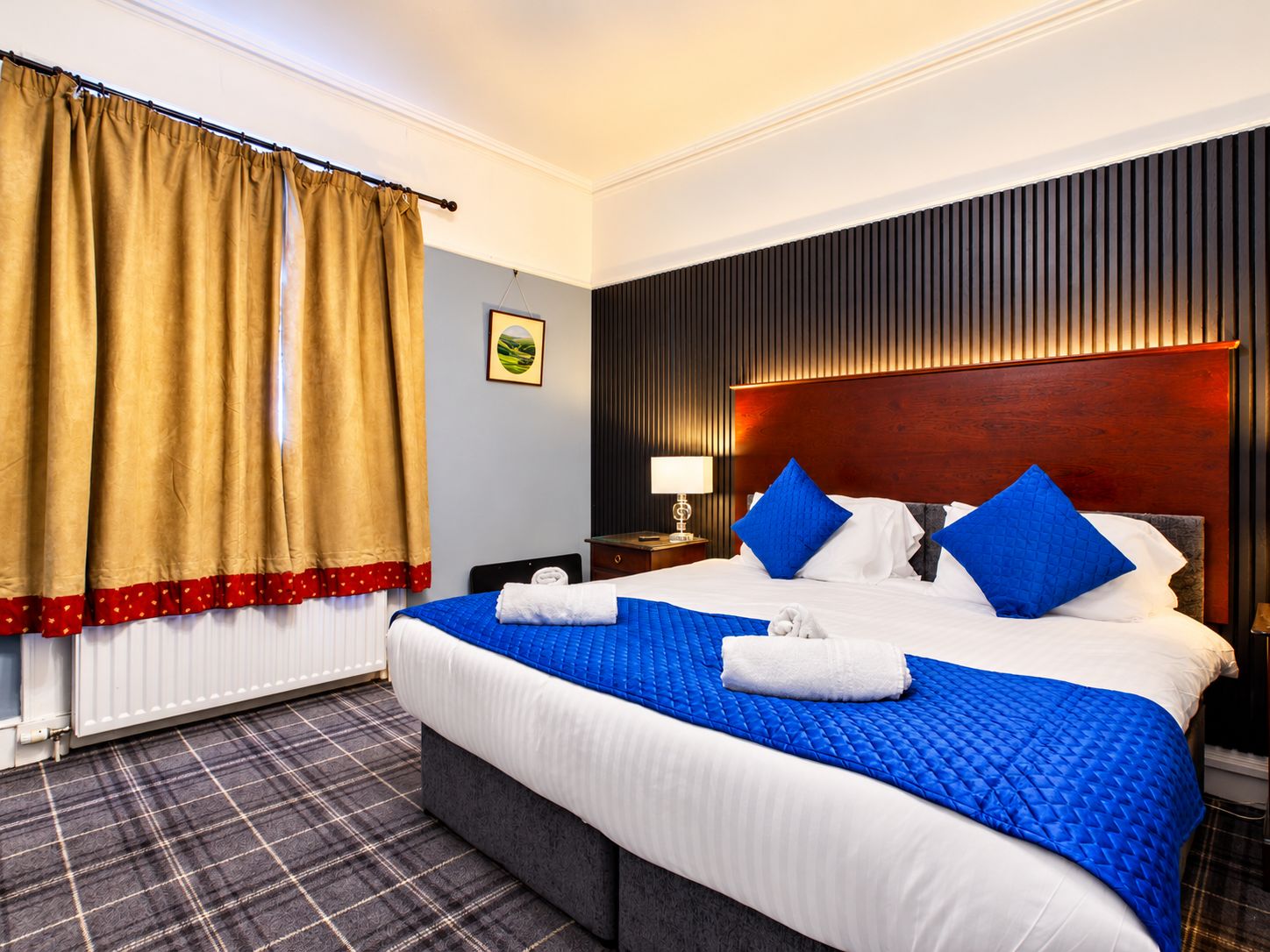 Habitación de hotel con una cama grande, una mesita de noche, una lámpara, un ventanal con cortinas y un radiador.