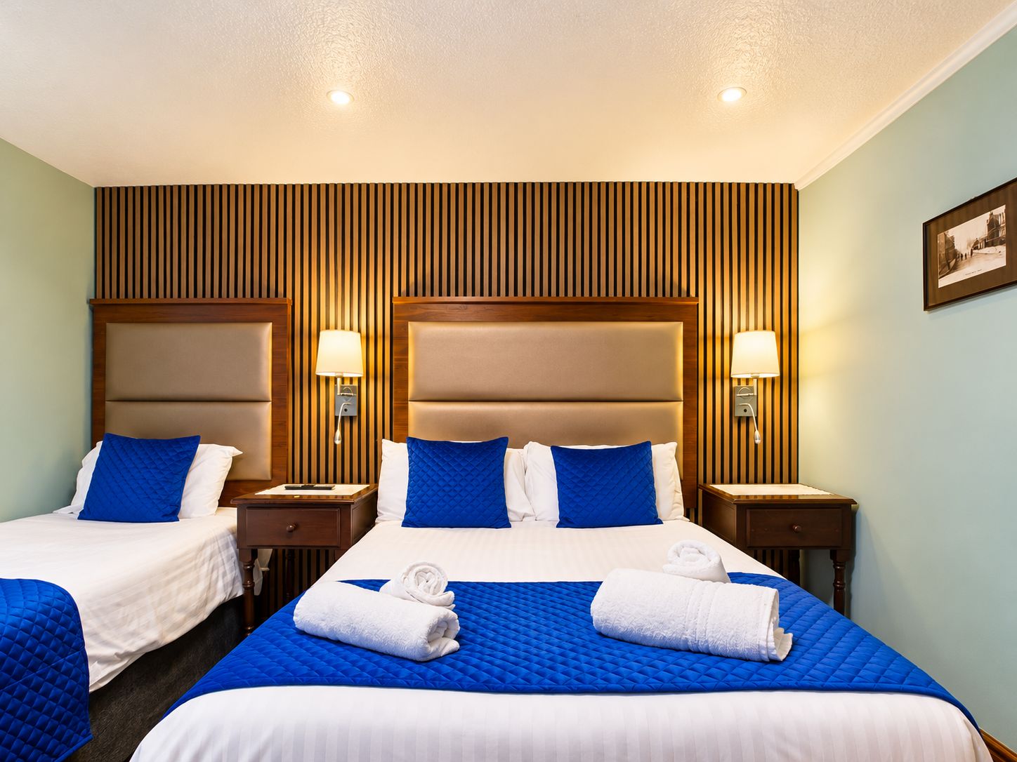Quarto de hotel com duas camas, cada uma com colcha azul acolchoada e almofadas brancas, com cabeceiras de madeira e abajures laterais.