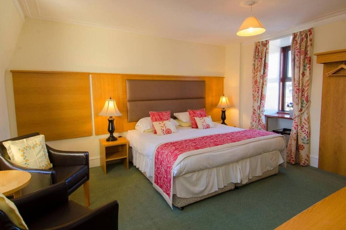 A hotel room with a large bed, two bedside lamps, a wooden headboard, a chair, a small table, and a window with curtains.