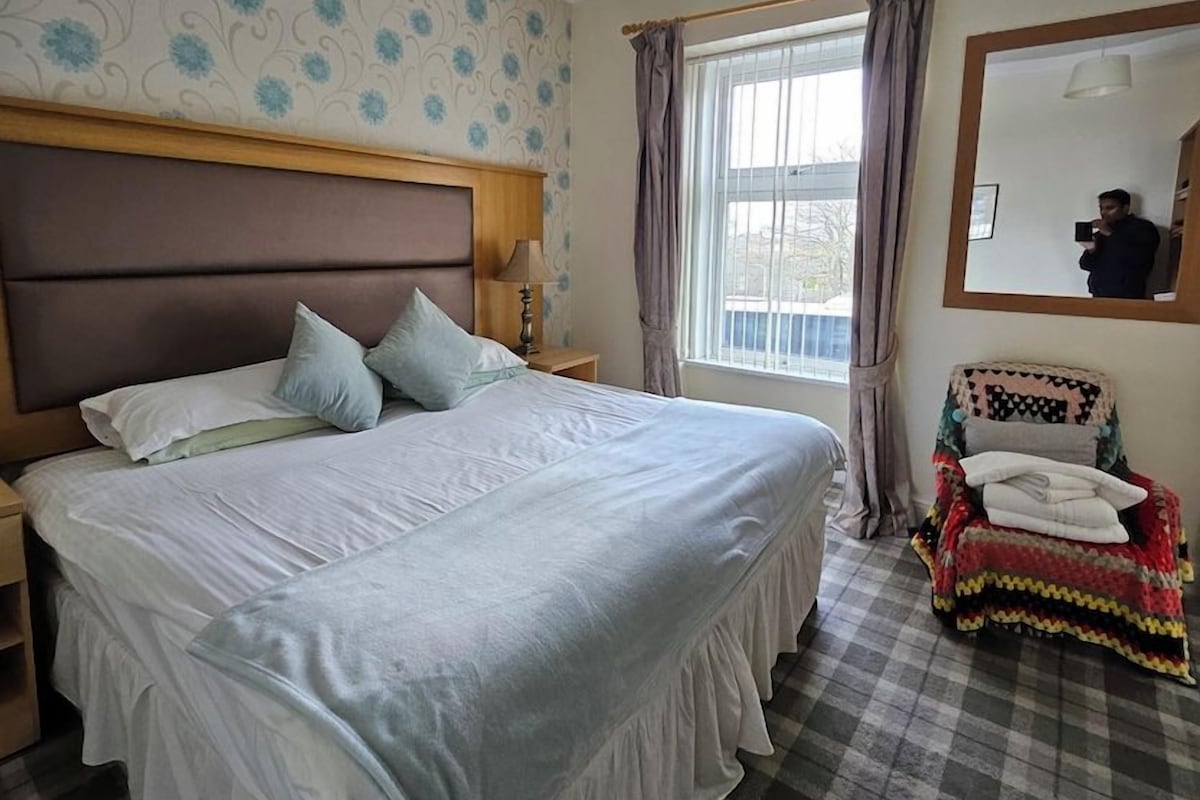Une chambre avec un grand lit, une chaise recouverte d’une couverture, une fenêtre avec des rideaux et un miroir au mur.