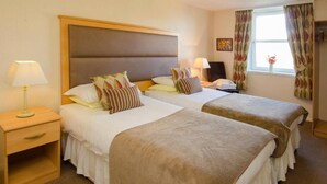 A hotel room with two beds, each with a headboard, bedside tables, lamps, and a window with curtains. - Homesly The Station Hotel (Banff)