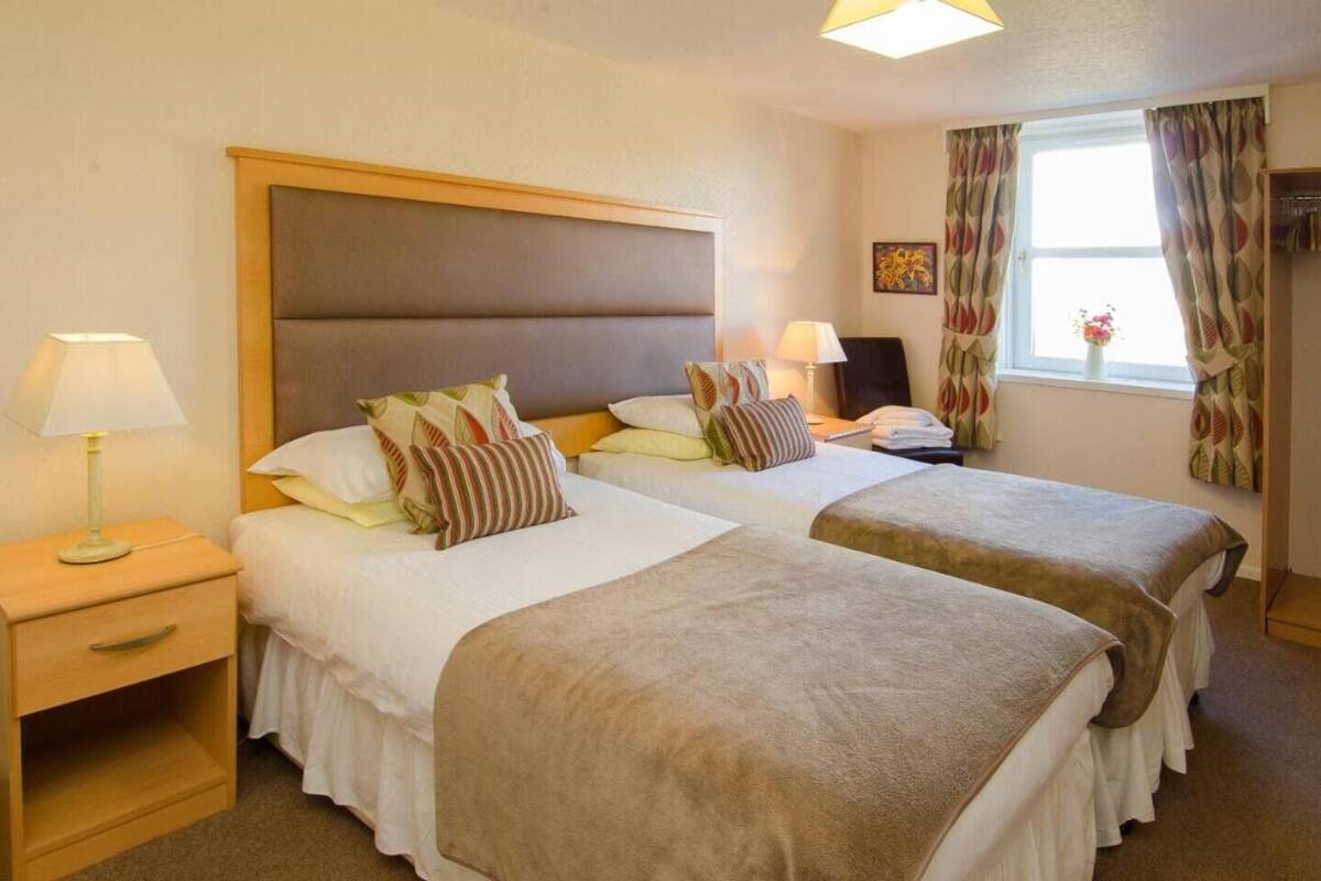 A hotel room with two beds, each with a headboard, bedside tables, lamps, and a window with curtains.