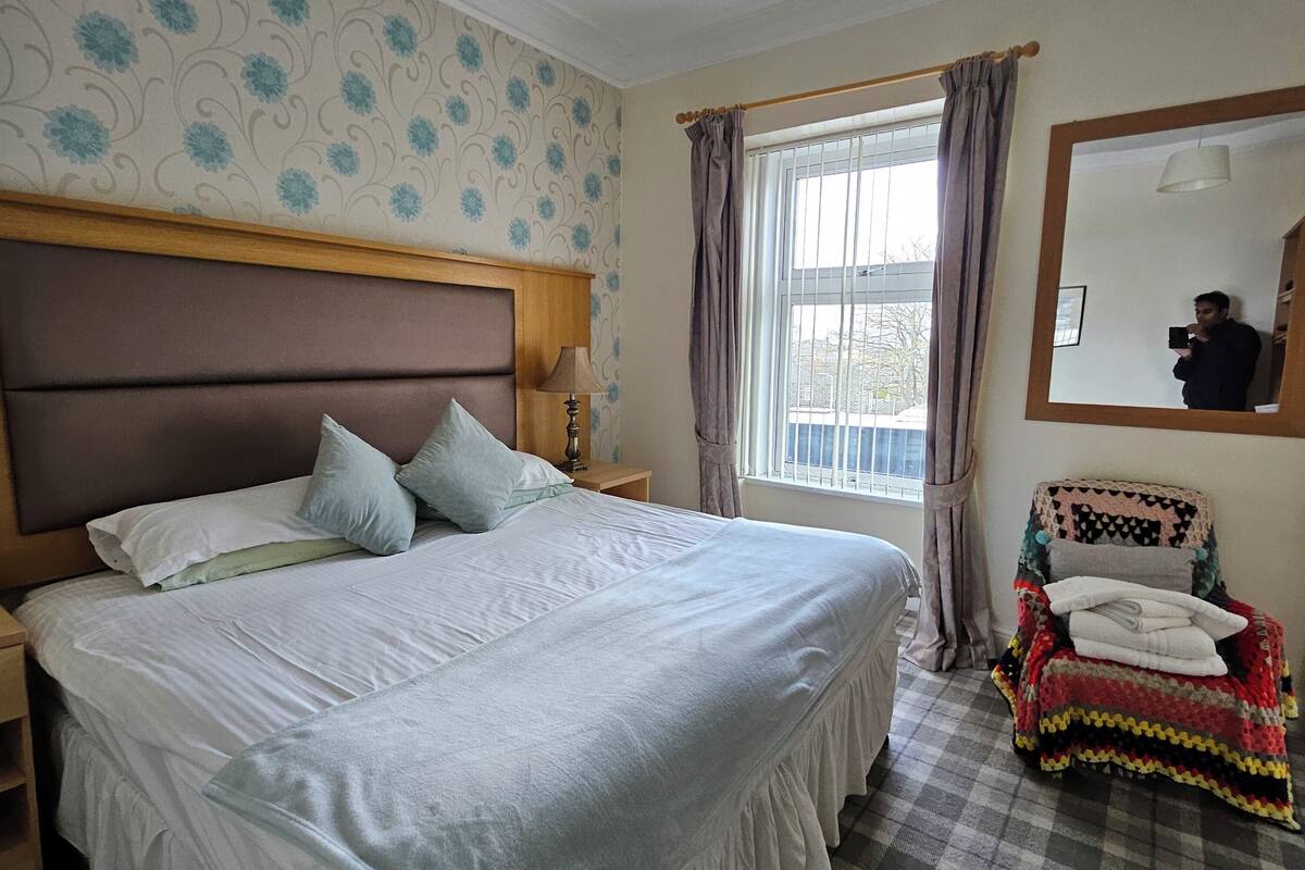 A neatly arranged bedroom with a large bed, a bedside table with a lamp, a chair with a blanket, and a window with curtains.