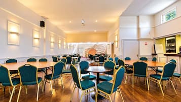 A hotel conference room with multiple tables and chairs, a kitchen area, and a view of the outdoors.