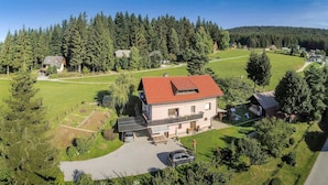 Una casa de dos pisos con techo rojo, rodeada por un césped cuidado y un bosque frondoso.