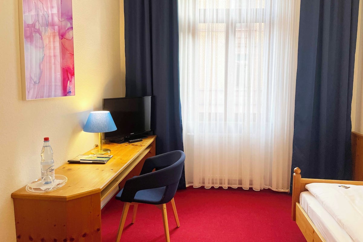 A hotel room with a wooden desk, a television, a chair, a bed, and a window with sheer curtains.