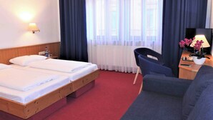 A hotel room with two single beds, a wooden headboard, a red carpet, a blue armchair, a desk with a lamp and flowers, and a window with sheer curtains. - Hotel Krokodil (Heidelberg)
