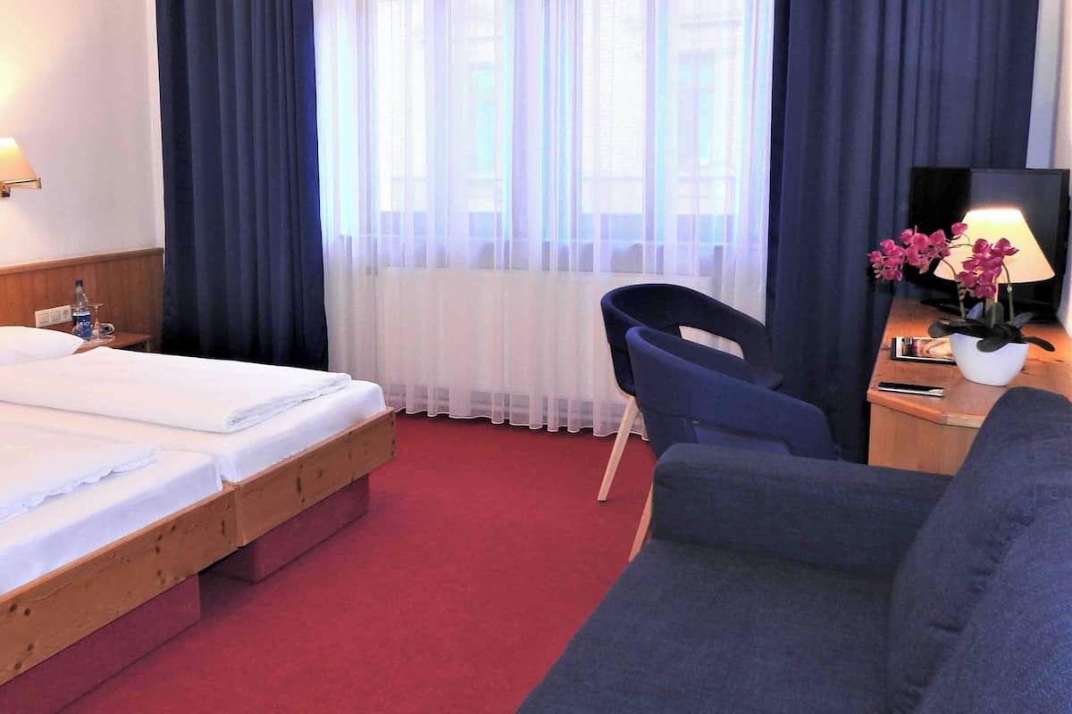 A hotel room with two single beds, a wooden headboard, a red carpet, a blue armchair, a desk with a lamp and flowers, and a window with sheer curtains.