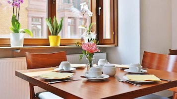 Mesa de comedor preparada para cuatro, con platos, tazas y cubiertos blancos, un jarrón con flores y plantas en macetas sobre un alféizar.